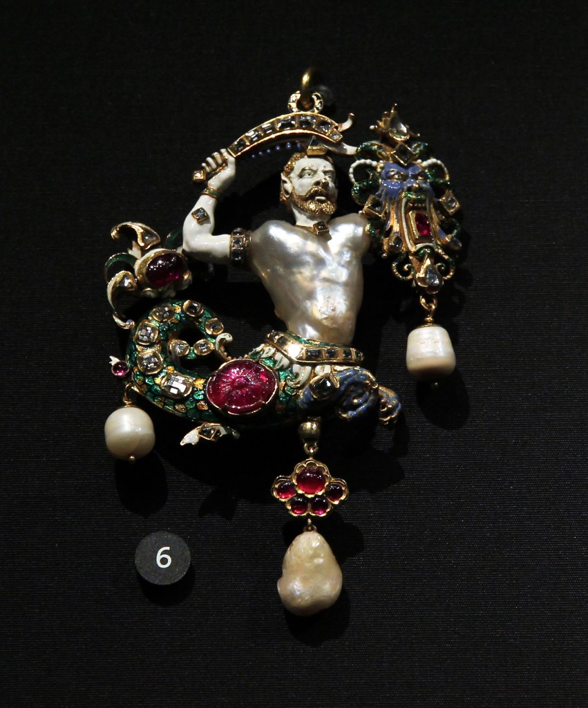 A fantastic image of a merman made of an irregular shaped pearl and gold with enamel, garnets, and rubies showing the merman holding the head of a dragon in one hand and a sword in the other
